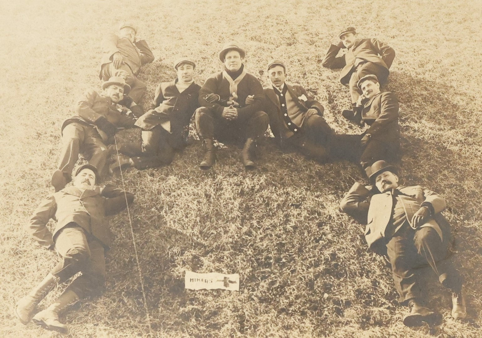 Lot 125 - Early Selfie, Antique Boarded Photograph Of A Group Of Men, Hikers, Forming An H On The Ground, With Hiker Flag On Grass, Note Man Left Center Pulling String To Operate The Shutter, Take Photo