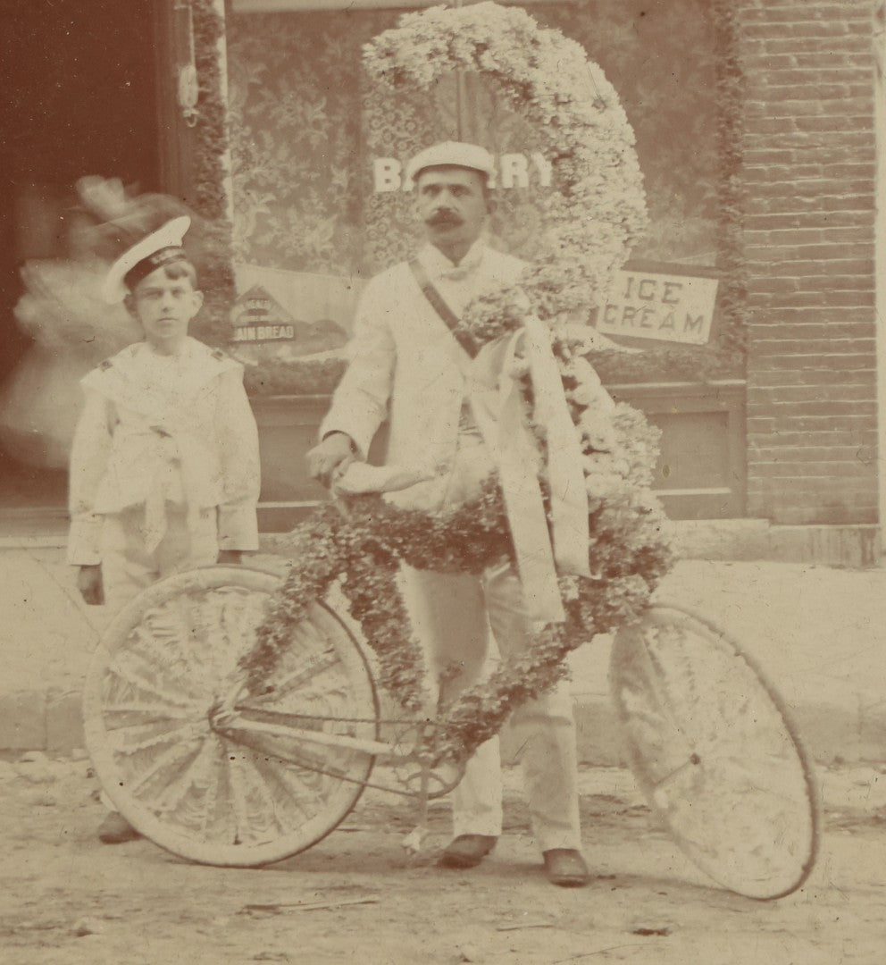 Lot 045 - Antique Framed Photo Of A Young Boy And Man, Likely His Father, With A Bike Covered In Flowers, Flower Moon, Boy In Naval Outfit, Likely For Parade, With Ice Cream Shop In Background