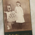 Lot 130 - Antique Cabinet Card Photograph Of Young Girl In White Dress Posing Next To Table With Photos In Picture, P.I.P., W.L. Fox Photographer, Battle Creek, Michigan, Identified As Anna May Downs