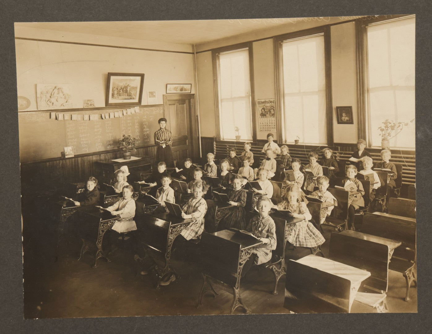 Lot 060 - Antique Boarded Photograph Of The Interior Of A 2Nd Grade Classroom With School Children, Teacher Holding Books,