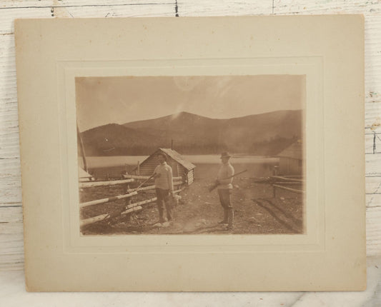Lot 097 - Antique Boarded Photograph Of Men Holding Rifles, Gamefowl, With Hunting Cabin, Lake, And Mountains In Background