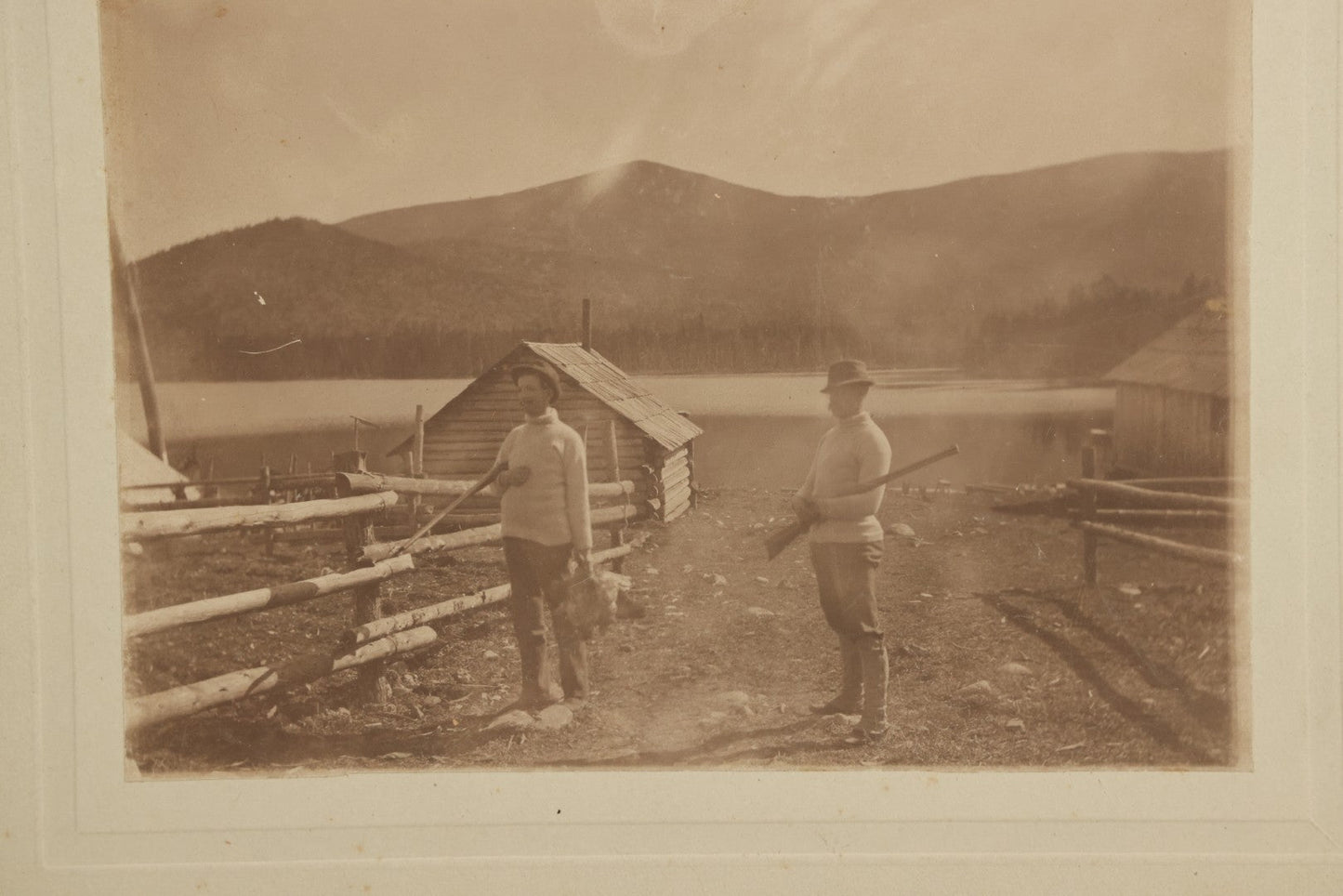 Lot 097 - Antique Boarded Photograph Of Men Holding Rifles, Gamefowl, With Hunting Cabin, Lake, And Mountains In Background
