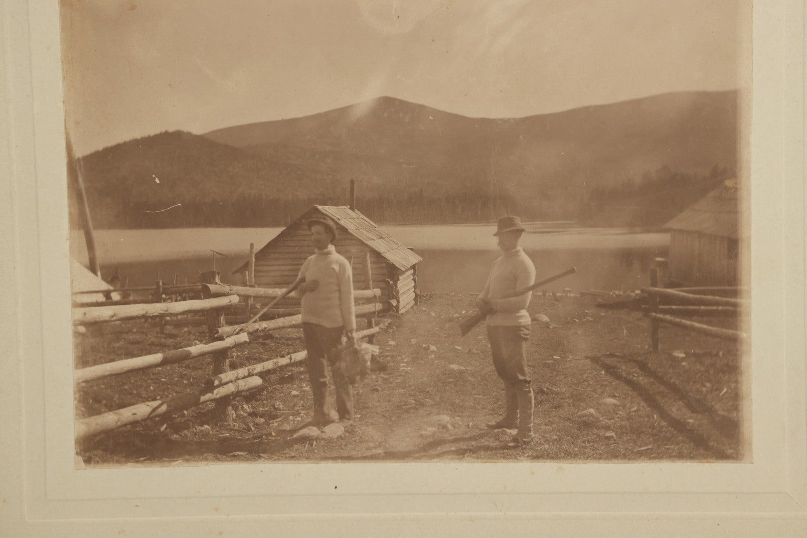 Lot 097 - Antique Boarded Photograph Of Men Holding Rifles, Gamefowl, With Hunting Cabin, Lake, And Mountains In Background