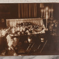 Lot 101 - Vintage Paper Funeral Photograph Of Young Man In Coffin Adorned With Floral Arrangements And "Dear Brother" Ribbon, Crosses, Photos, Candles And Religious Art Visible, Including Photo Of WWI Soldier In Top Left