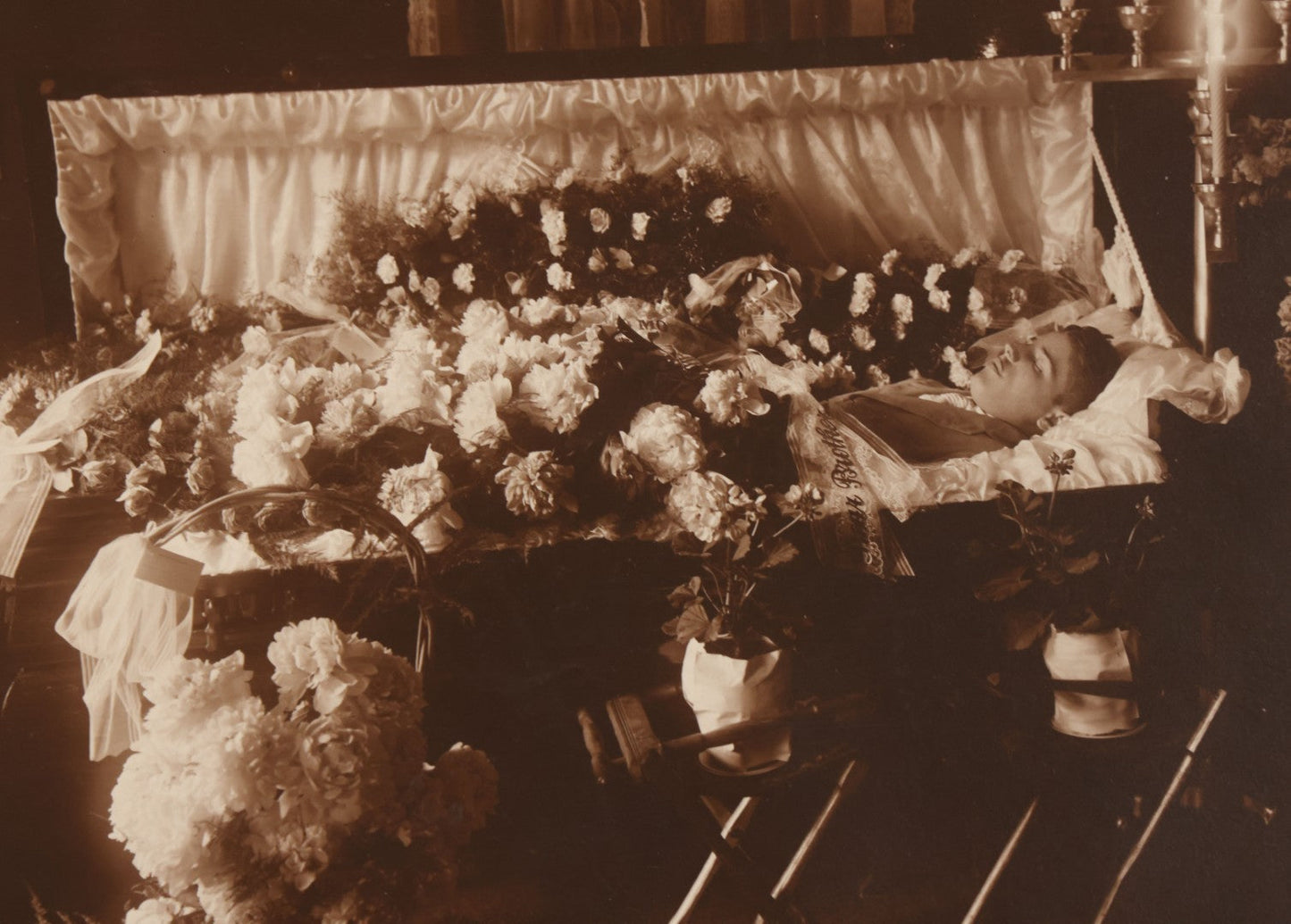Lot 101 - Vintage Paper Funeral Photograph Of Young Man In Coffin Adorned With Floral Arrangements And "Dear Brother" Ribbon, Crosses, Photos, Candles And Religious Art Visible, Including Photo Of WWI Soldier In Top Left