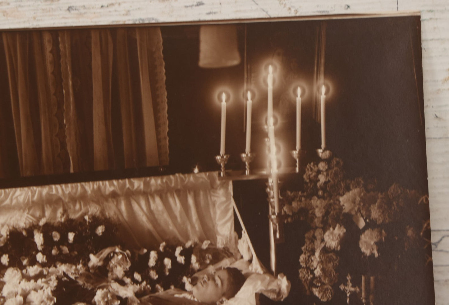 Lot 101 - Vintage Paper Funeral Photograph Of Young Man In Coffin Adorned With Floral Arrangements And "Dear Brother" Ribbon, Crosses, Photos, Candles And Religious Art Visible, Including Photo Of WWI Soldier In Top Left