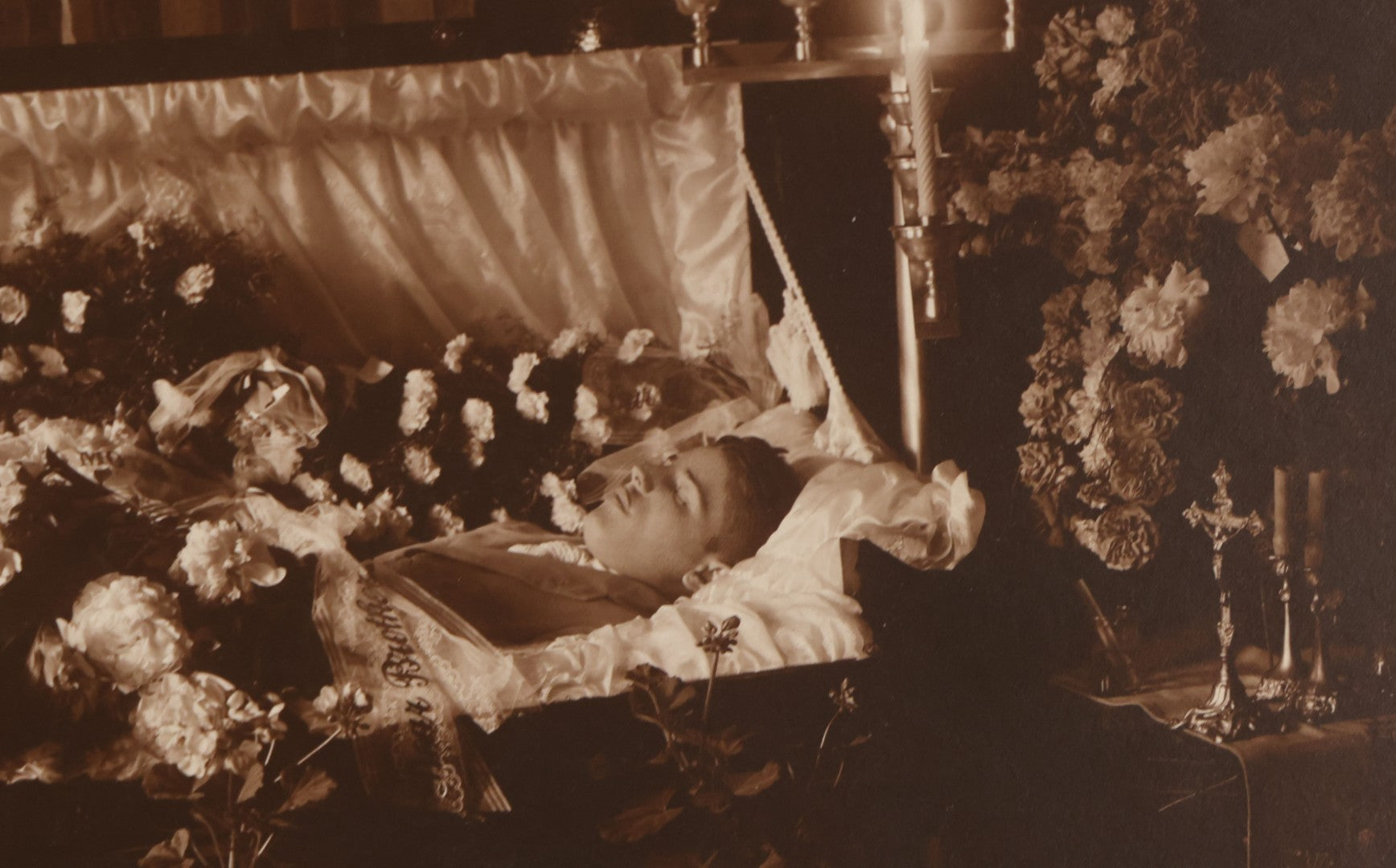Lot 101 - Vintage Paper Funeral Photograph Of Young Man In Coffin Adorned With Floral Arrangements And "Dear Brother" Ribbon, Crosses, Photos, Candles And Religious Art Visible, Including Photo Of WWI Soldier In Top Left