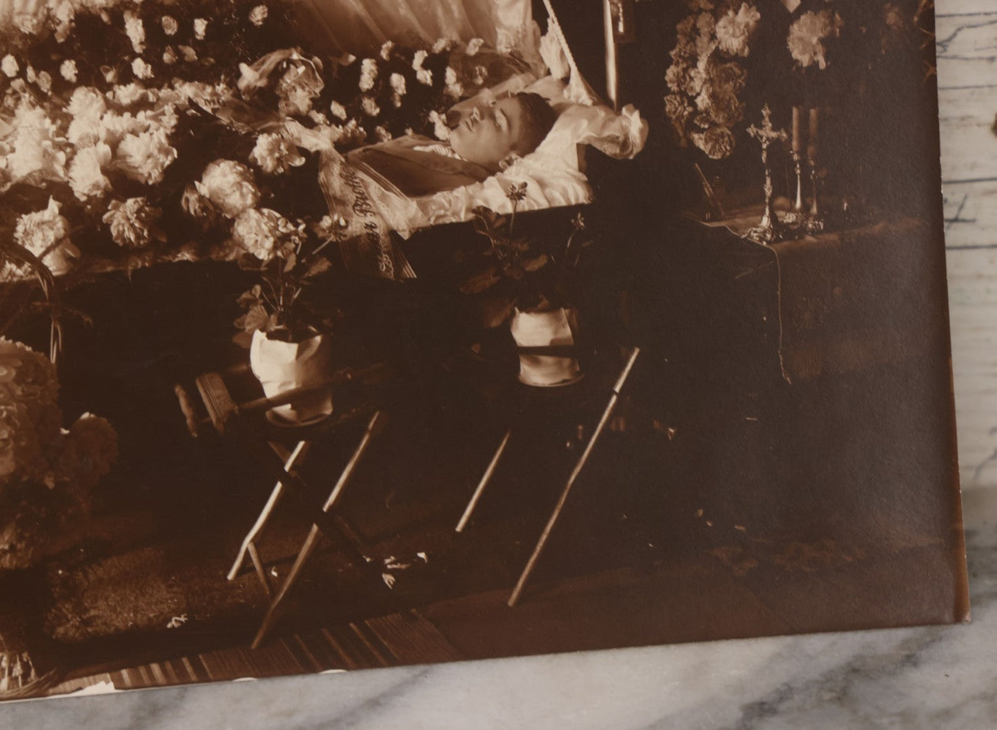 Lot 101 - Vintage Paper Funeral Photograph Of Young Man In Coffin Adorned With Floral Arrangements And "Dear Brother" Ribbon, Crosses, Photos, Candles And Religious Art Visible, Including Photo Of WWI Soldier In Top Left