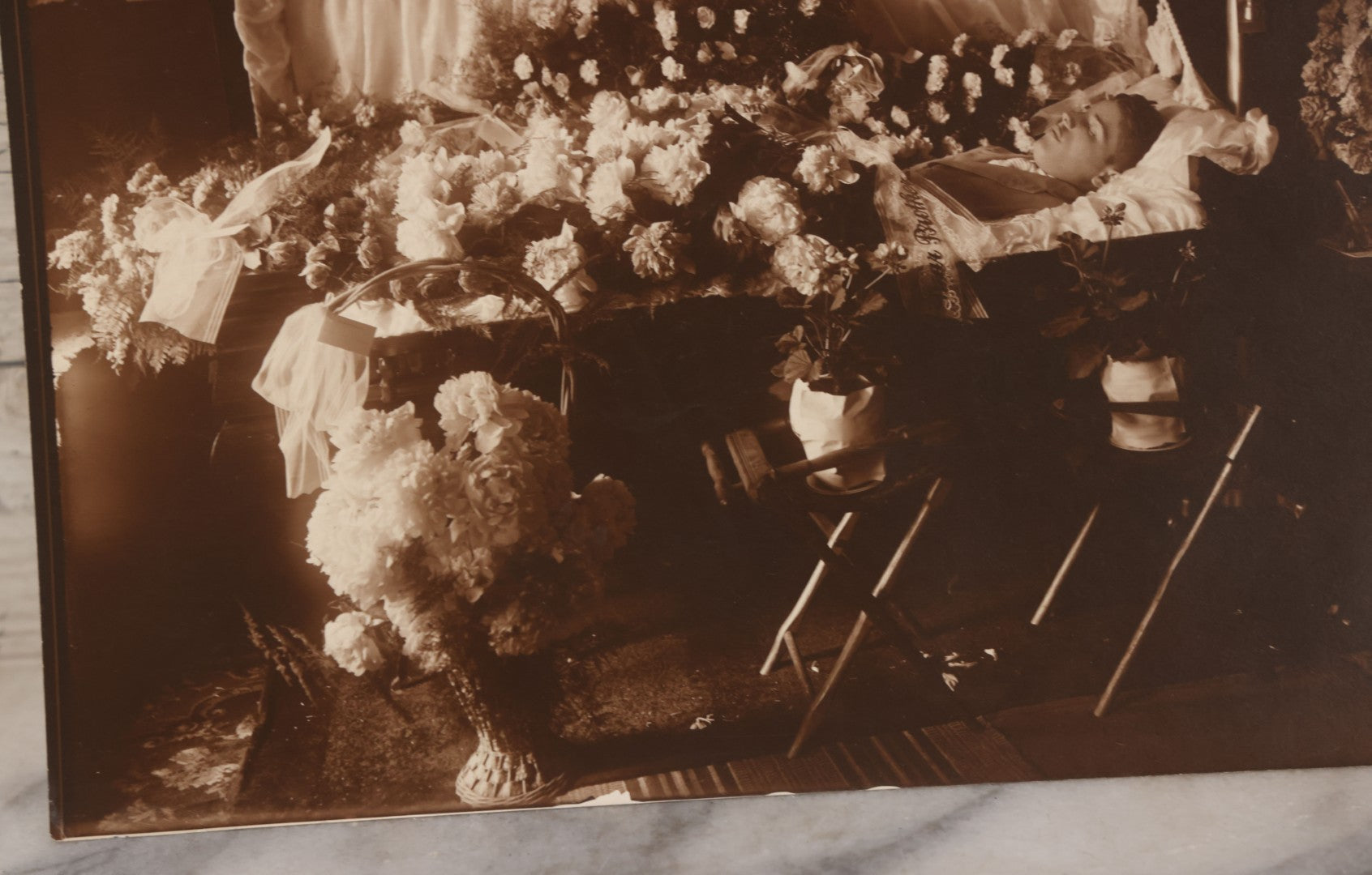 Lot 101 - Vintage Paper Funeral Photograph Of Young Man In Coffin Adorned With Floral Arrangements And "Dear Brother" Ribbon, Crosses, Photos, Candles And Religious Art Visible, Including Photo Of WWI Soldier In Top Left