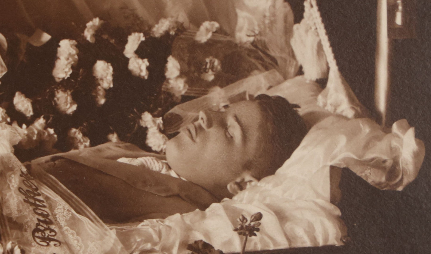 Lot 101 - Vintage Paper Funeral Photograph Of Young Man In Coffin Adorned With Floral Arrangements And "Dear Brother" Ribbon, Crosses, Photos, Candles And Religious Art Visible, Including Photo Of WWI Soldier In Top Left