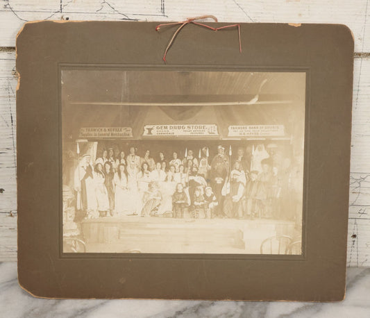 Lot 125 - Antique Boarded Photograph Of Cast Of Biblical Themed Play Onstage In Costume, With Advertisements Above Stage For Gem Drug Store, Farmers Bank, And Trawick & Neville Suppliers Of Merchandise, Likely From Souris, North Dakota