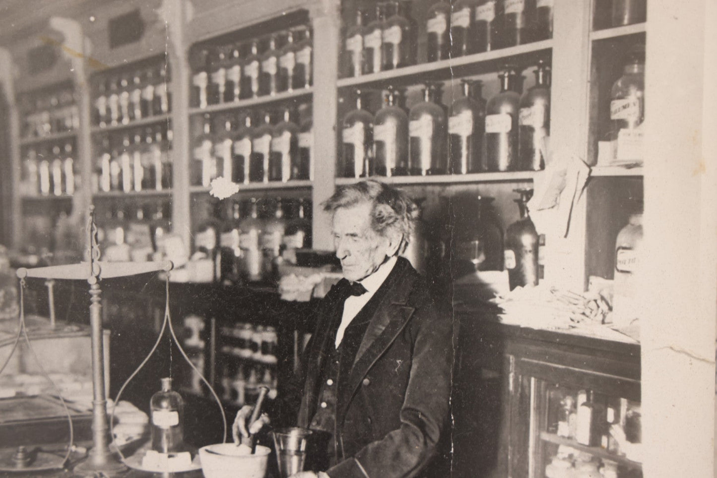 Lot 093 - Antique Large Format Photograph Of Elderly Pharmacist Compounding Medicine At The Pharmacy Counter, With Many Apothecary Bottles And Tools Visible