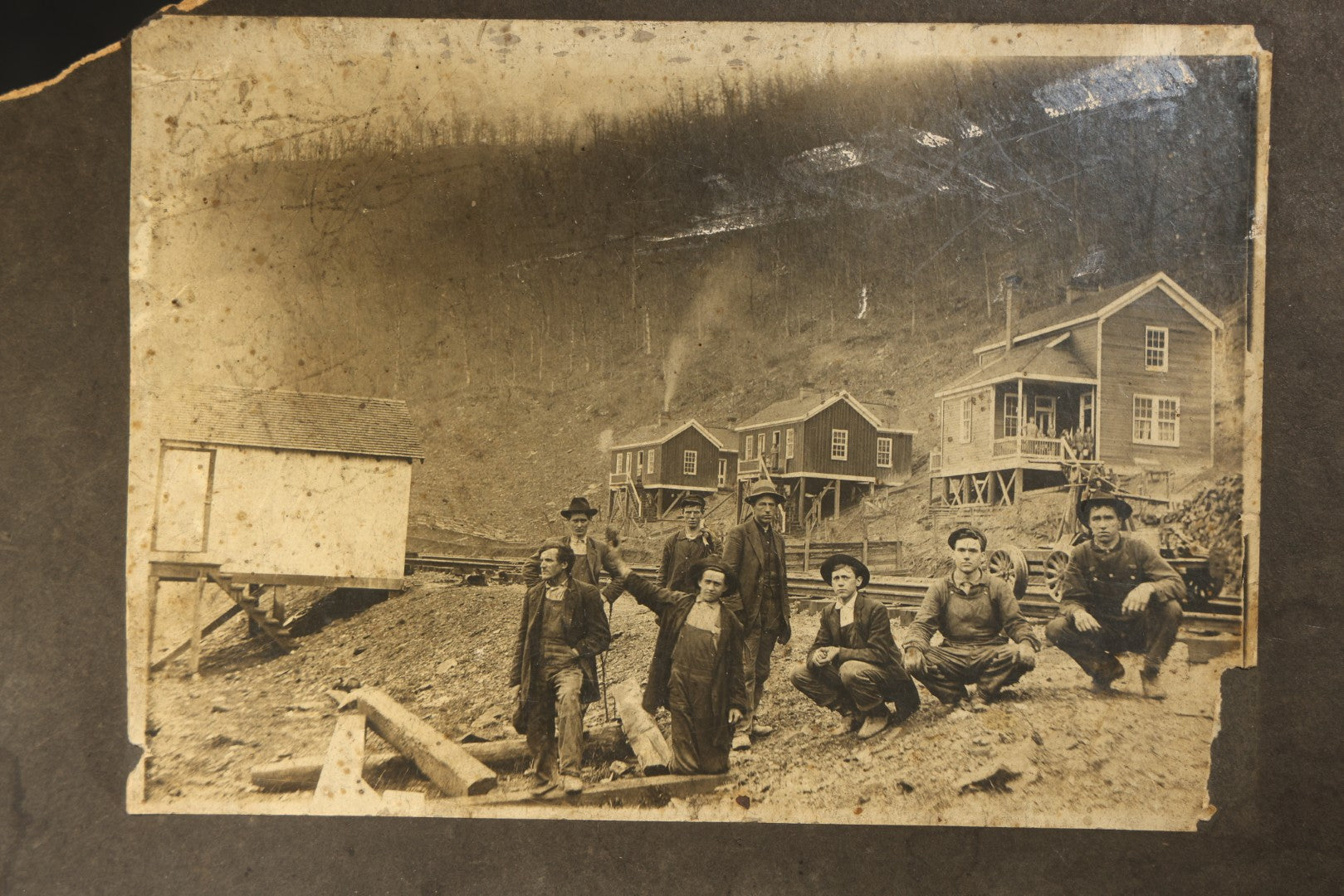 Antique Board-Mounted Occupational Photograph of Eight Railroad Workers Posed Over Railroad Ties at a Hillside Camp with Frame Buildings