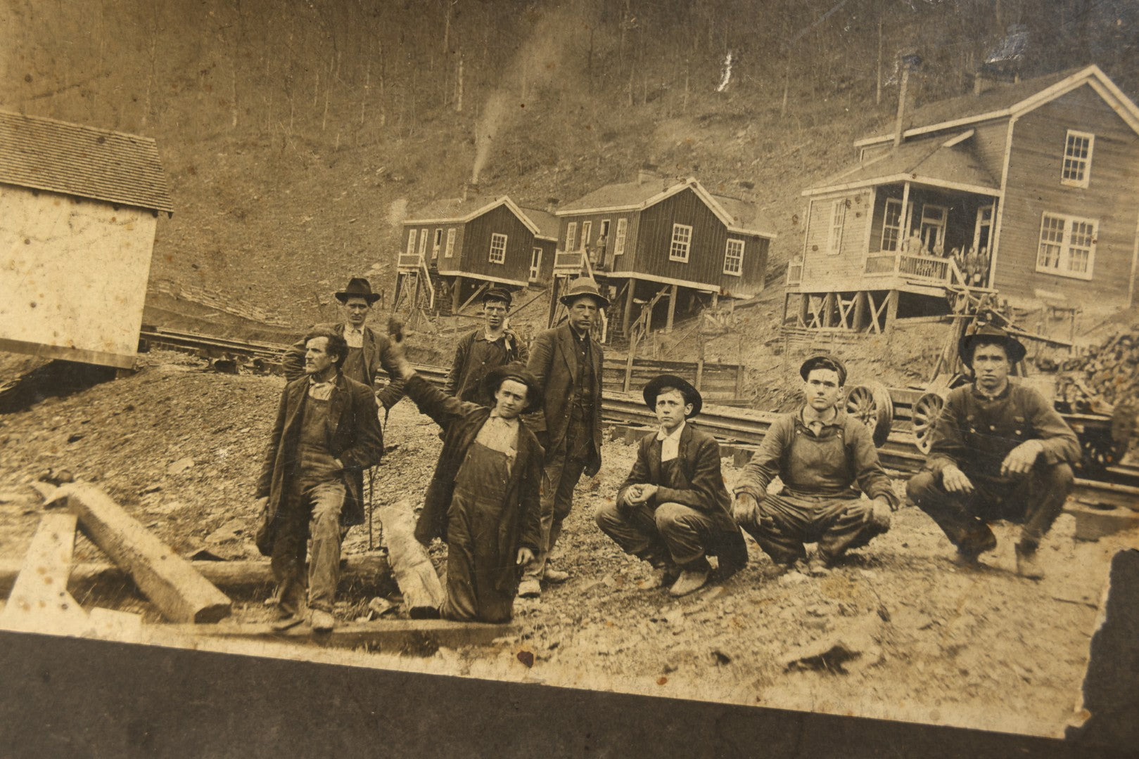 Antique Board-Mounted Occupational Photograph of Eight Railroad Workers Posed Over Railroad Ties at a Hillside Camp with Frame Buildings