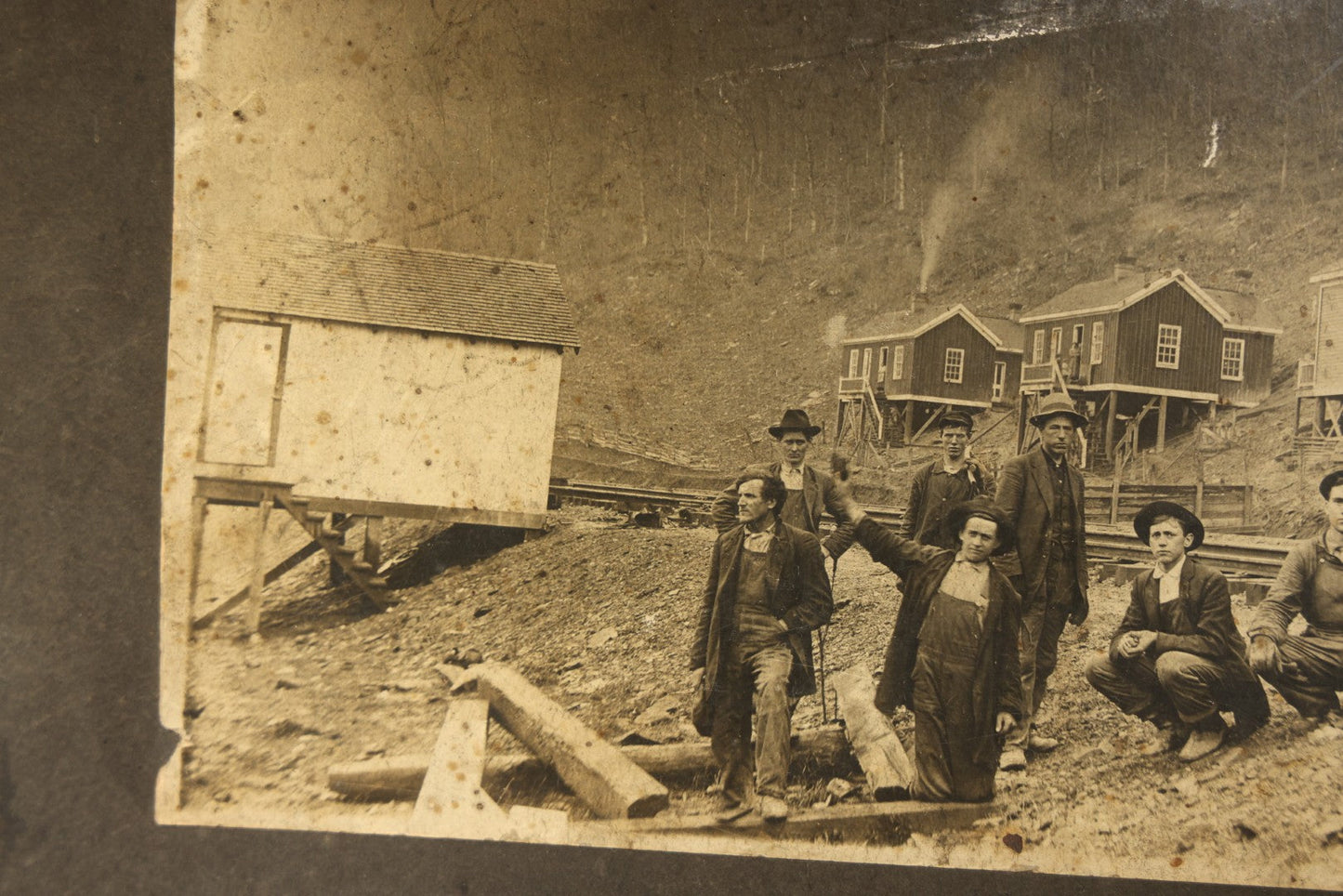 Antique Board-Mounted Occupational Photograph of Eight Railroad Workers Posed Over Railroad Ties at a Hillside Camp with Frame Buildings