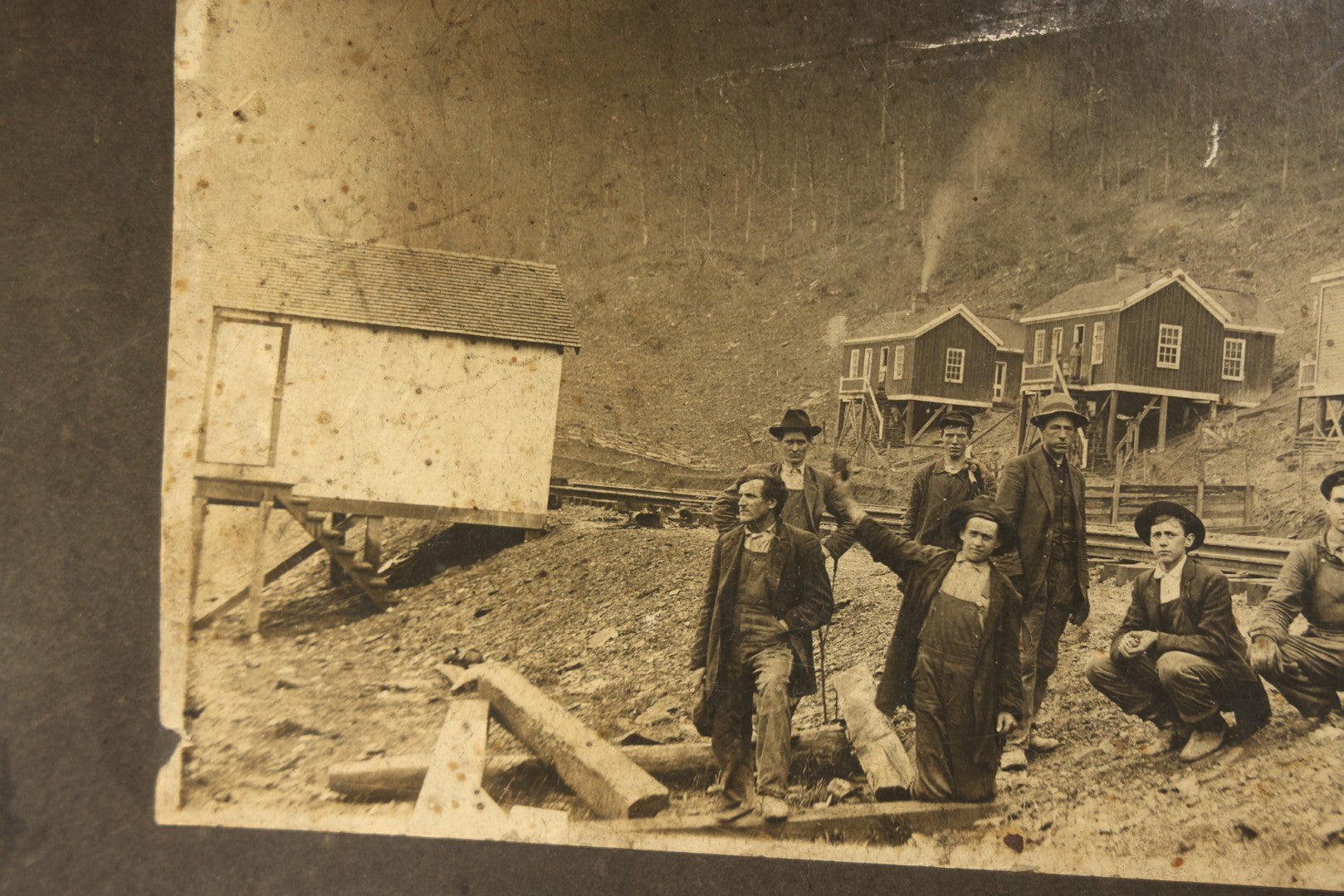 Antique Board-Mounted Occupational Photograph of Eight Railroad Workers Posed Over Railroad Ties at a Hillside Camp with Frame Buildings