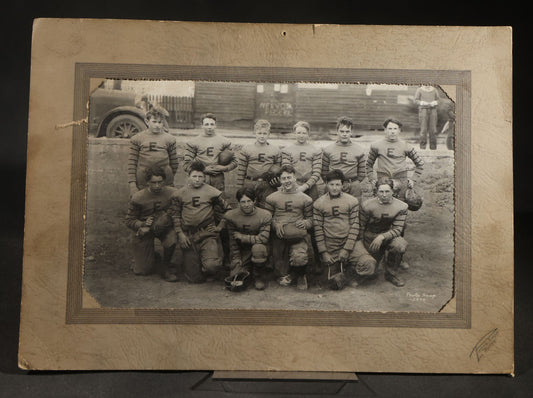 Antique Board-Mounted Photograph of a Football Team in "E" Letter Jerseys with Striped Sleeves, 12 Players with Leather Helmets and Footballs, Photographed by Photo Shop, Butte, Montana, Circa 1920s
