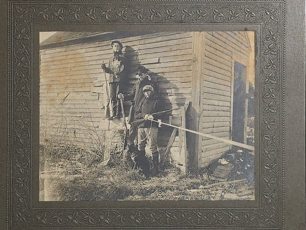 Antique Board-Mounted "Selfie" Photograph of Three Hunters with Rifles and Game Posed Outside a Wooden Building, Self-Timed Shutter Release