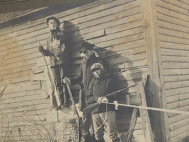 Antique Board-Mounted "Selfie" Photograph of Three Hunters with Rifles and Game Posed Outside a Wooden Building, Self-Timed Shutter Release