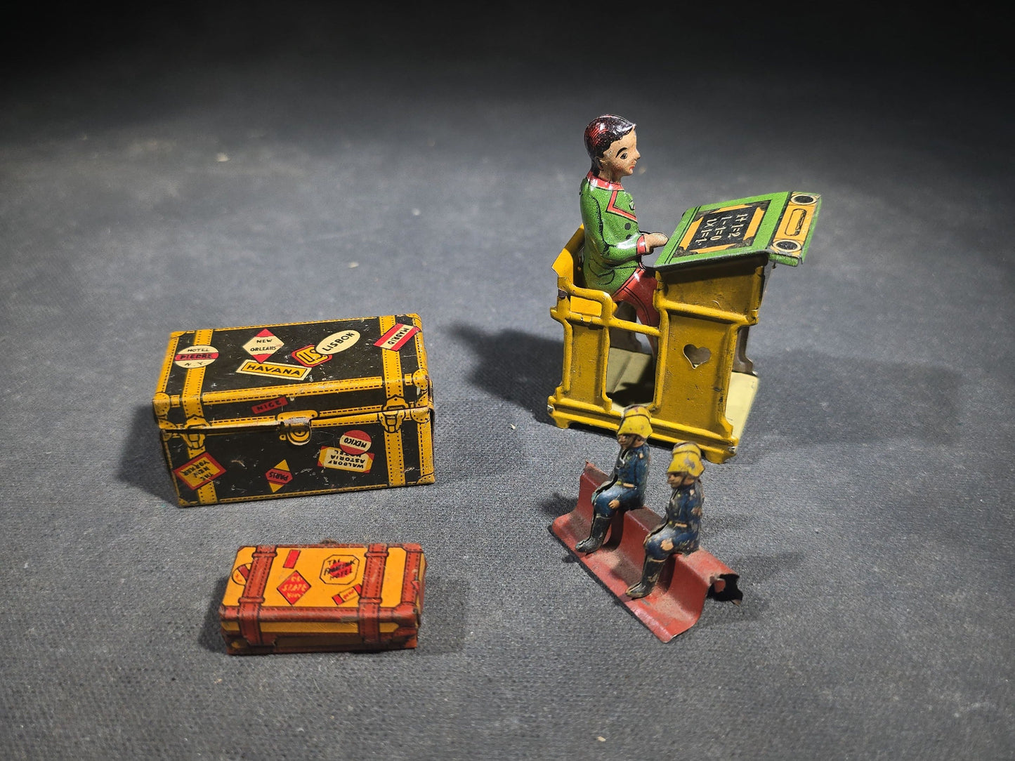 Grouping of Four Antique and Vintage Tin Lithograph Miniature Toys including Student at Desk, Two Miniature Travel Suitcases with Hotel and Travel Labels, and Pair of Seated Figures in Helmets